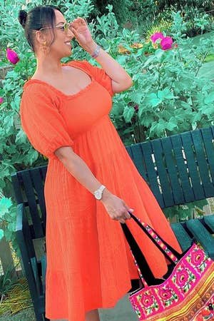 a reviewer in a casual orange dress holding a bag with vibrant patterns, standing by a bench in a garden setting