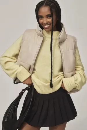 Person wearing a beige puffy vest, light sweater, black pleated skirt, and holding a black bag, smiling confidently