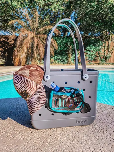 A beach bag by a pool holds a zebra-print cap, sunglasses, and sunscreen